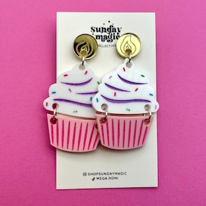 Puede incluir: Un par de pendientes de cupcake rosa y blanco con glaseado morado y blanco y chispas de colores. Los pendientes est&aacute;n unidos a ganchos de pendientes dorados con un dise&ntilde;o de llama.