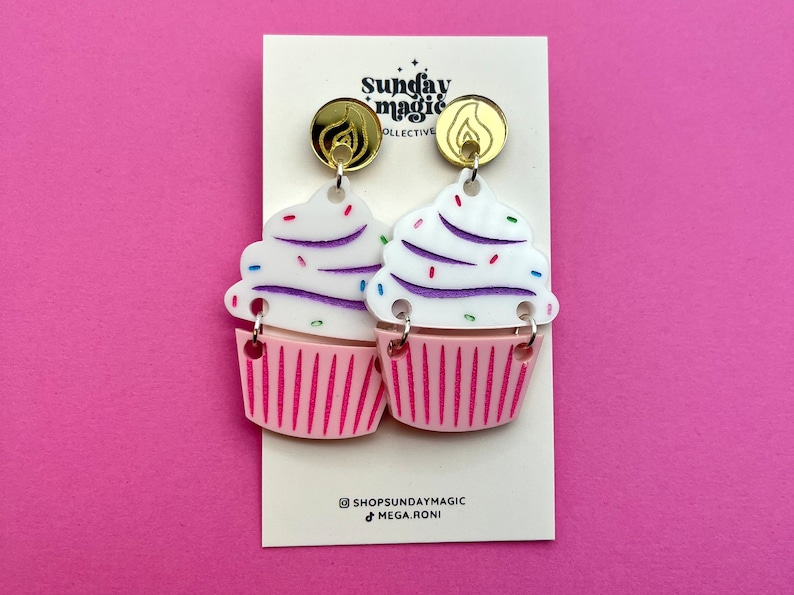 Puede incluir: Un par de pendientes de cupcake rosa y blanco con glaseado morado y blanco y chispas de colores. Los pendientes est&aacute;n unidos a ganchos de pendientes dorados con un dise&ntilde;o de llama.