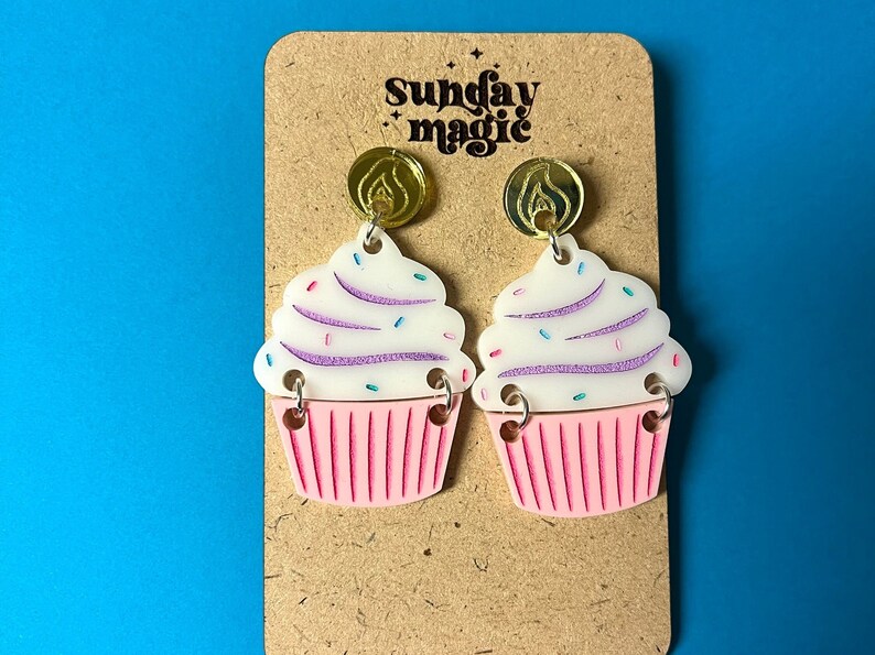 Puede incluir: Un par de pendientes con forma de cupcake con glaseado rosa y blanco, chispas y detalles dorados. Los pendientes est&aacute;n en una tarjeta marr&oacute;n con el texto "Sunday Magic".