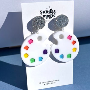 May include: A pair of white paint palette earrings with colorful paint drops. The earrings are attached to silver glitter studs.