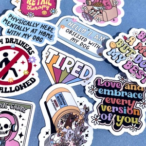 May include: A collection of colorful stickers with various phrases and designs. Some of the stickers include "Energy Drainers Not Allowed", "Bit of a Mess But Doin My Best", "Love and Embrace Every Version of You", "True Crime", "Tired", "Hello I Am Obsessed With My Dog", and "Physically Here Mentally At Home With My Dog".
