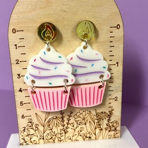 Puede incluir: Un par de pendientes con forma de cupcake con glaseado rosa y blanco, chispas moradas y blancas y detalles dorados. Los pendientes se muestran en una regla de madera con un dise&ntilde;o floral.