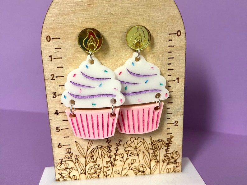Puede incluir: Un par de pendientes con forma de cupcake con glaseado rosa y blanco, chispas moradas y blancas y detalles dorados. Los pendientes se muestran en una regla de madera con un dise&ntilde;o floral.
