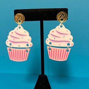 Puede incluir: Un par de pendientes con forma de cupcake con glaseado rosa y blanco y espolvoreado. Los pendientes est&aacute;n hechos de acr&iacute;lico y tienen ganchos de color dorado.