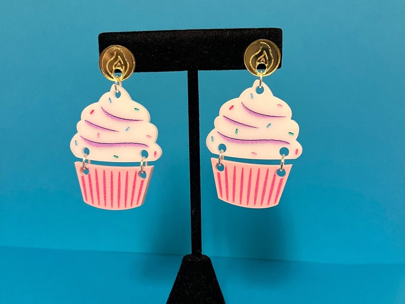 Puede incluir: Un par de pendientes con forma de cupcake con glaseado rosa y blanco y espolvoreado. Los pendientes est&aacute;n hechos de acr&iacute;lico y tienen ganchos de color dorado.
