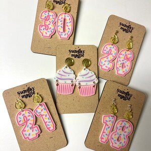 Puede incluir: Cinco pares de pendientes con tem&aacute;tica de cumplea&ntilde;os en tarjetas de exhibici&oacute;n. Los pendientes est&aacute;n hechos de acr&iacute;lico rosa y presentan un cupcake, los n&uacute;meros 18, 21, 26 y 30, con chispas de colores y adornos de velas doradas. Las tarjetas son marrones y tienen el texto "Sunday Magic" impreso en ellas.