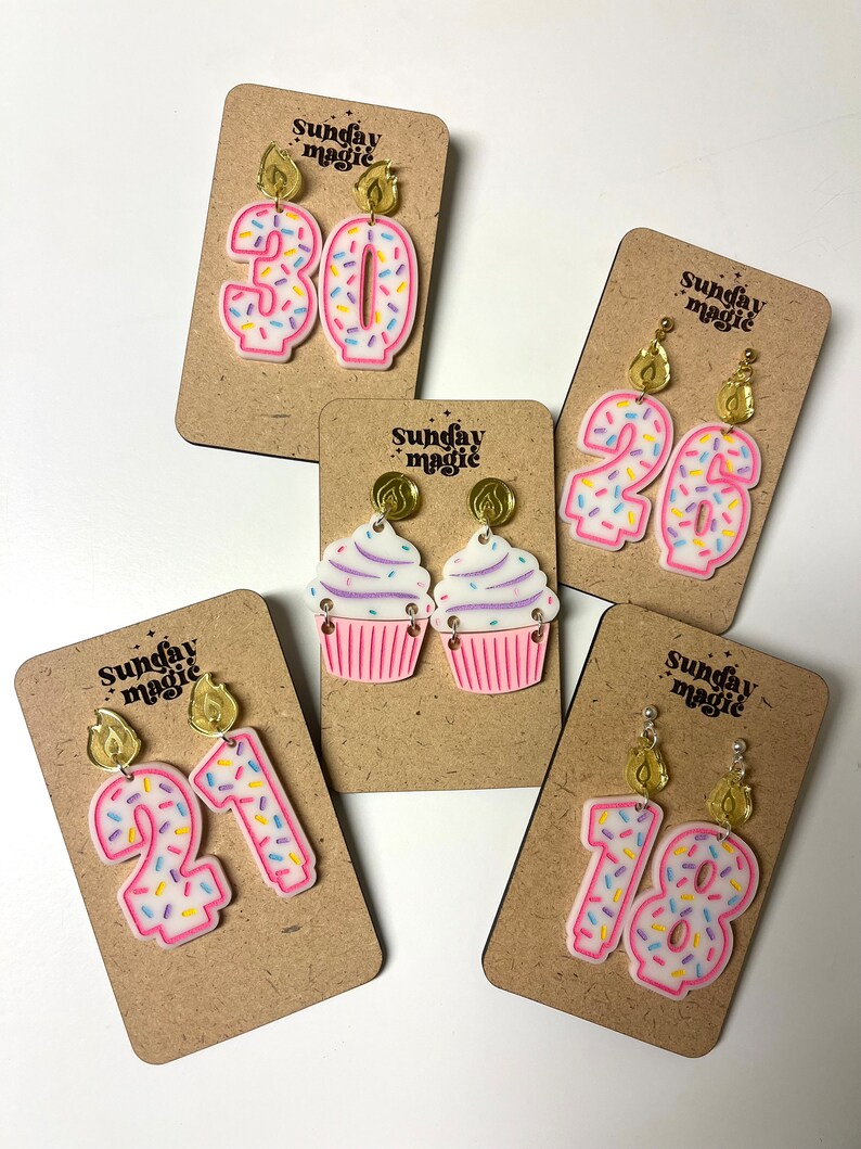Puede incluir: Cinco pares de pendientes con tem&aacute;tica de cumplea&ntilde;os en tarjetas de exhibici&oacute;n. Los pendientes est&aacute;n hechos de acr&iacute;lico rosa y presentan un cupcake, los n&uacute;meros 18, 21, 26 y 30, con chispas de colores y adornos de velas doradas. Las tarjetas son marrones y tienen el texto "Sunday Magic" impreso en ellas.