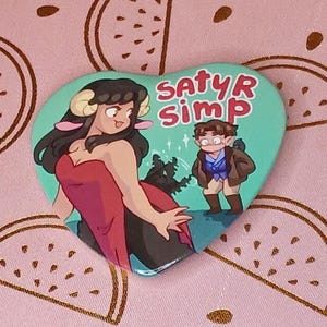 May include: A heart-shaped button with a green background features a cartoon illustration of a woman with horns and a red dress, and a man in a blue jacket and brown pants. The text "SATYR SIMP" is written in pink letters on the button.