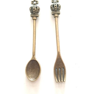 May include: A set of two miniature spoons with crown-shaped handles. One spoon is a spoon and the other is a fork. Both are made of a gold-toned metal.
