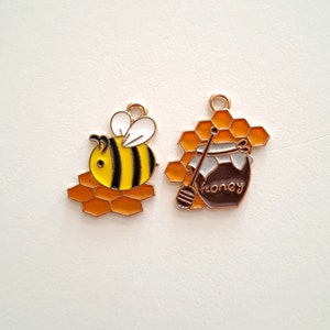 Peut inclure: Deux breloques en métal doré représentant une abeille de dessin animé sur un nid d'abeilles et un pot de miel avec un bâton de miel sur un nid d'abeilles.