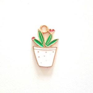 Puede incluir: Encanto de metal dorado con una planta en maceta con hojas verdes y flores rosas. La maceta es blanca con detalles dorados y tiene un pequeño agujero en la parte superior para sujetarlo a una cadena o pulsera.
