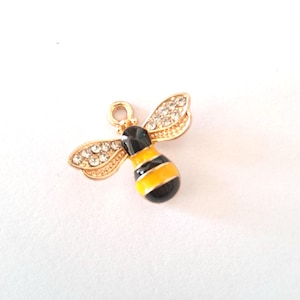 May include: A gold-coloured bee charm with black and yellow stripes on the body. The wings are embellished with small, clear crystals. The charm has a loop at the top for attaching to jewellery or other crafts.