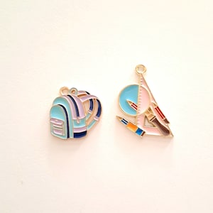 May include: Two gold-coloured metal charms, one shaped like a blue backpack with purple and black accents, and the other shaped like a pink triangle with a compass, ruler, and pencils.