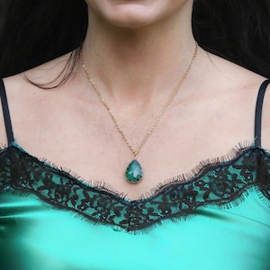 May include: A woman wearing a green satin top with black lace trim and a gold chain necklace with a large emerald green teardrop pendant.