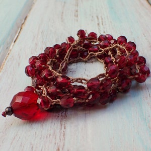 May include: A handmade red beaded bracelet with a large, faceted pendant. The beads are a deep red color and are strung on a tan cord. The bracelet is displayed on a distressed, light blue and white wooden surface.
