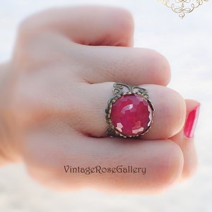 Może przedstawiać: Pierścionek z dużym, fasetowanym, czerwonym kamieniem szlachetnym osadzonym w ozdobnej, złoconej oprawie. Pierścionek jest na palcu.