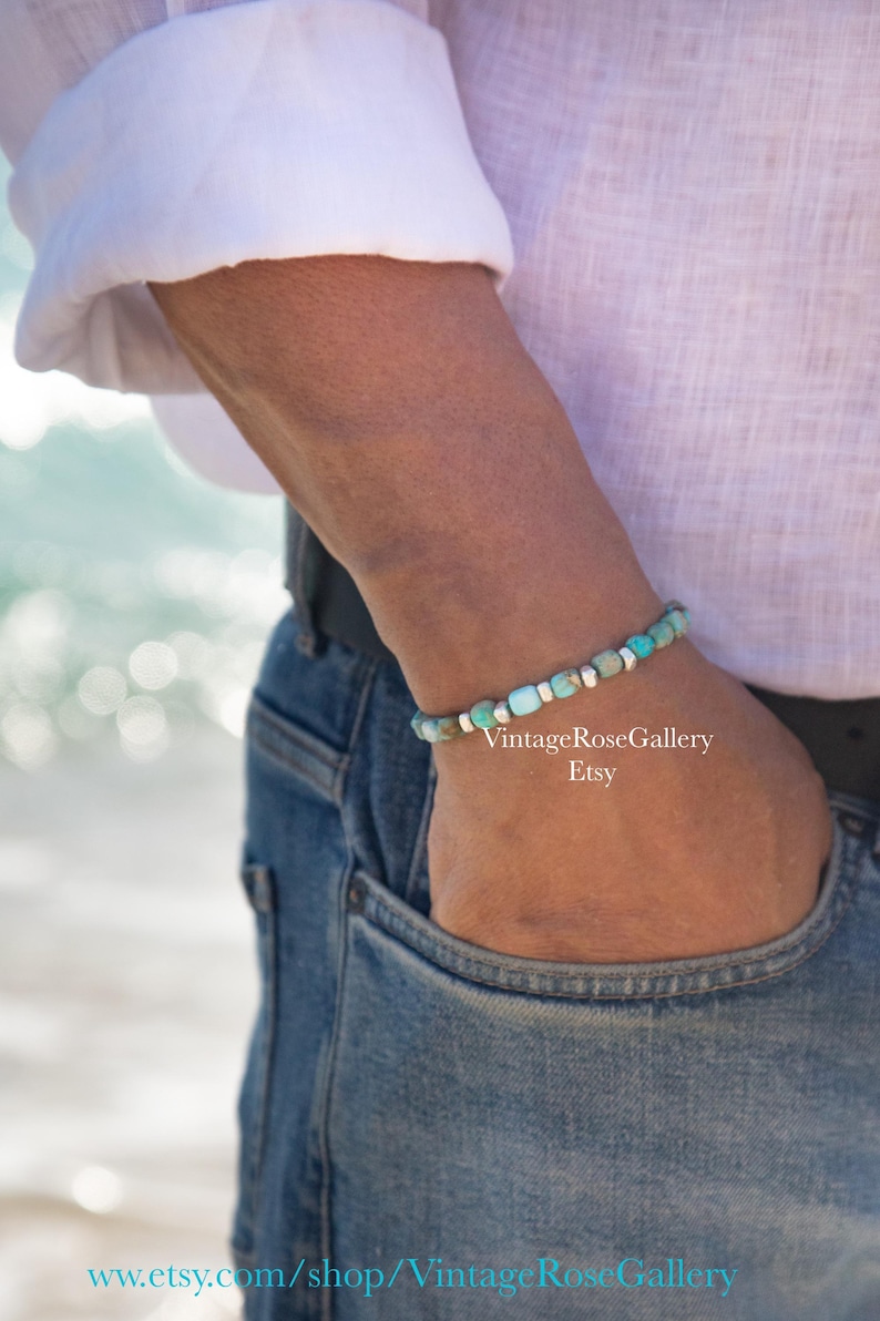 May include: A beaded bracelet featuring small, cylindrical turquoise-colored stones interspersed with tiny, polished silver-toned metal spacers. The bracelet is worn on a wrist, showcasing a repeating pattern of blue-green beads and metallic accents.