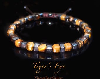 May include: A bracelet featuring tiger's eye and dark grey beads. The tiger's eye beads are a warm orange-brown colour, and the dark grey beads are square-shaped. The bracelet is on a black surface. The text "Tiger's Eye" and "Vintage Rose Gallery" are in the image.