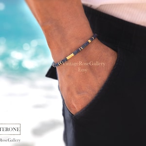 Puede incluir: Una pulsera de cuentas azul marino y dorada. La pulsera est&aacute; en una mu&ntilde;eca, con la mano en un bolsillo. El fondo es una imagen borrosa del oc&eacute;ano. El texto "TESTOSTERONE" est&aacute; en la parte inferior de la imagen.