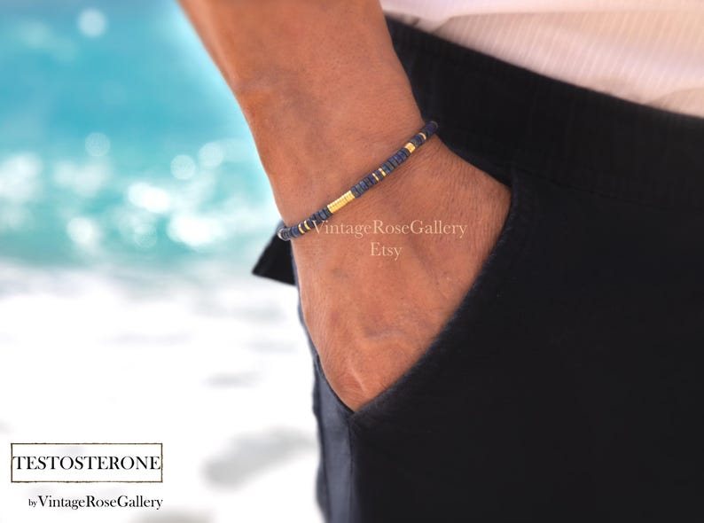 Puede incluir: Una pulsera de cuentas azul marino y dorada. La pulsera est&aacute; en una mu&ntilde;eca, con la mano en un bolsillo. El fondo es una imagen borrosa del oc&eacute;ano. El texto "TESTOSTERONE" est&aacute; en la parte inferior de la imagen.