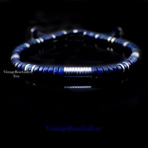 Puede incluir: Una pulsera azul oscuro con detalles plateados. La pulsera presenta peque&ntilde;as cuentas cuadradas azules y cuentas cil&iacute;ndricas plateadas. La pulsera se muestra sobre un fondo negro. El texto "VintageRoseGallery Etsy" es visible.