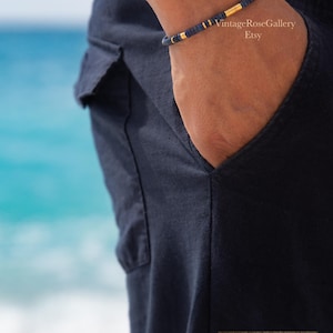 Puede incluir: Una pulsera de cuentas azul marino con detalles dorados. La pulsera se lleva en la mu&ntilde;eca, con un bolsillo azul marino en el fondo. El oc&eacute;ano es visible. El texto "TESTOSTERONE" est&aacute; en la parte inferior.