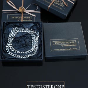 May include: A silver chain necklace presented in a black gift box, labelled "TESTOSTERONE" and "by VintageRoseGallery". The box is adorned with a gold ribbon and a gold flower. The background is black.