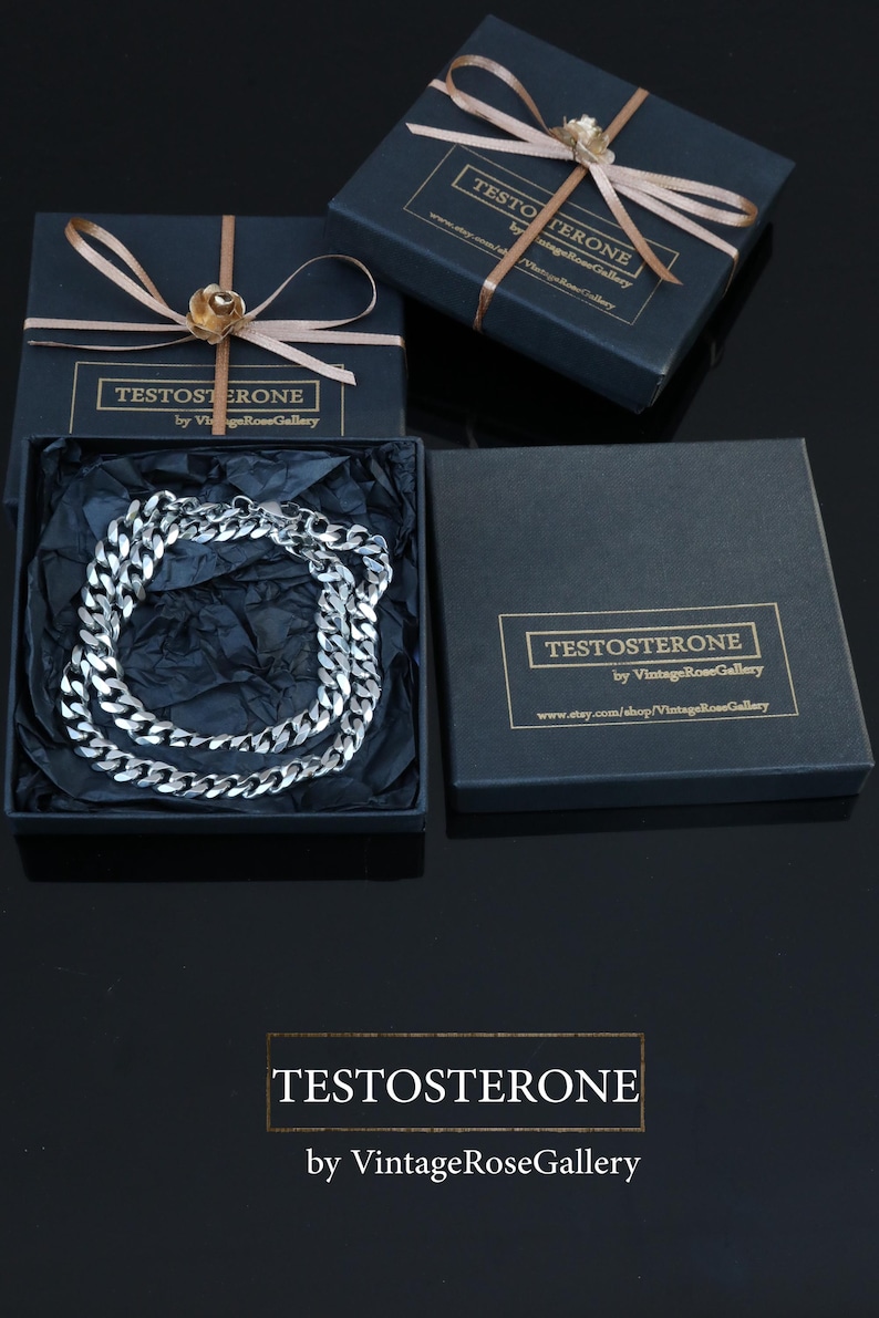May include: A silver chain necklace presented in a black gift box, labelled "TESTOSTERONE" and "by VintageRoseGallery". The box is adorned with a gold ribbon and a gold flower. The background is black.