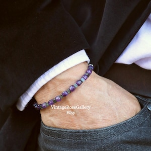 May include: A bracelet with purple gemstones and silver-coloured metal beads. The bracelet is worn on a wrist, visible against a white shirt cuff and a black jacket. The Etsy shop name is visible.