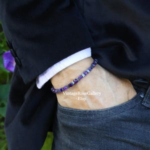 May include: A bracelet featuring purple and silver beads. The bracelet is worn on a wrist, with a black blazer and white shirt cuff visible. The person is wearing dark wash jeans. The Etsy shop name "VintageRoseGallery" is visible.