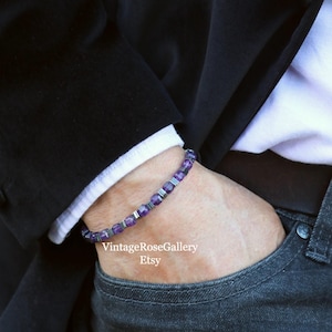 May include: A bracelet featuring purple and silver beads. The bracelet is worn on a wrist, with a black blazer and white shirt cuff visible. The Etsy shop name is visible in the image.