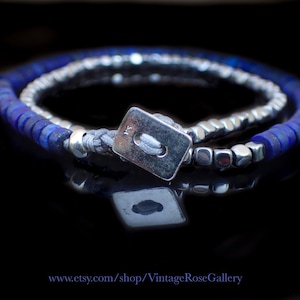 Könnte beinhalten: Ein Wickelarmband mit kleinen, flachen, kreisförmigen blauen Lapislazuli-Perlen und metallischen silberfarbenen Würfelperlen. Das Armband wird mit einem rechteckigen silberfarbenen Knopfverschluss an einer grauen Kordel geschlossen.