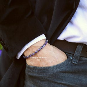 May include: A bracelet with purple and silver beads. The bracelet is worn on a wrist, with a black blazer and a white shirt visible in the background. The person is wearing dark wash jeans.