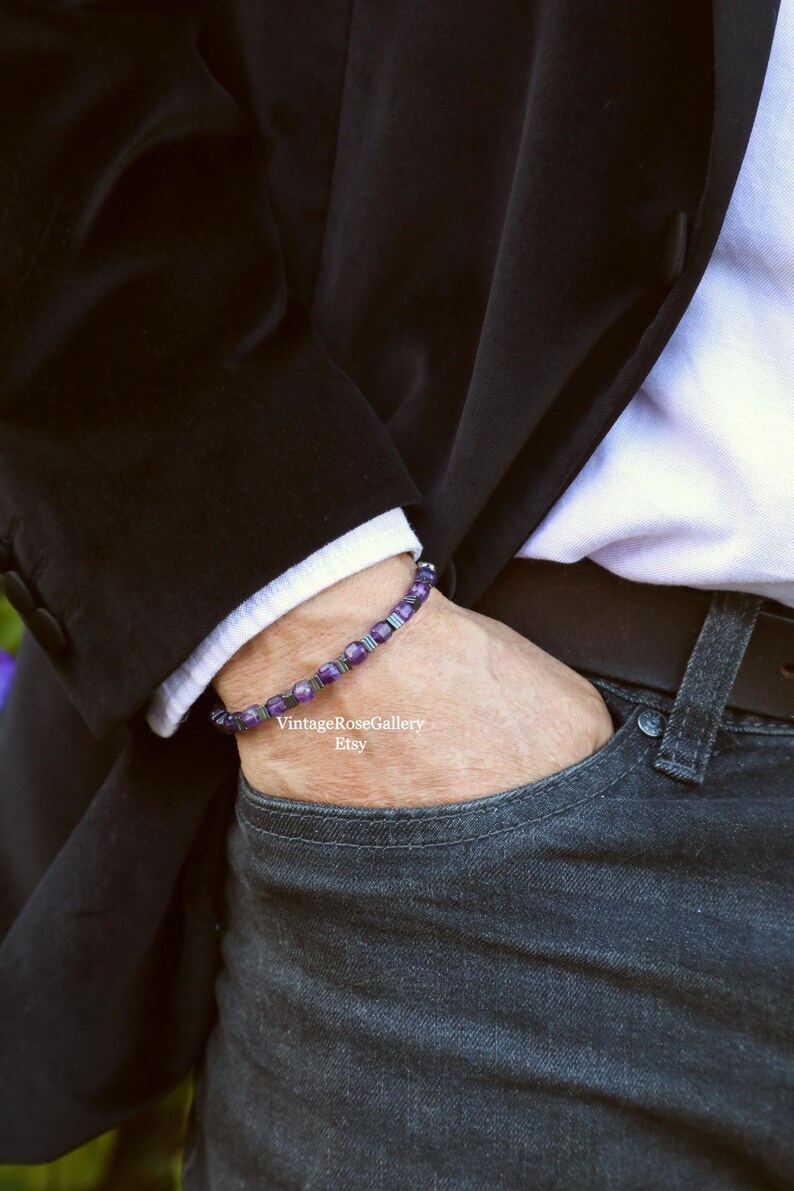 May include: A bracelet with purple and silver beads. The bracelet is worn on a wrist, with a black blazer and a white shirt visible in the background. The person is wearing dark wash jeans.