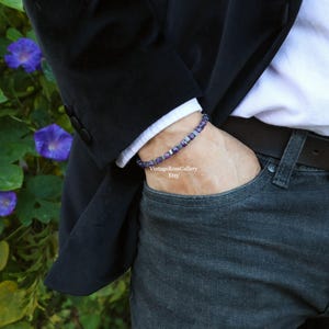 May include: A bracelet with purple and silver beads is worn on a wrist. The person is wearing a white shirt, a black blazer, and blue jeans. The background includes green foliage and purple flowers.