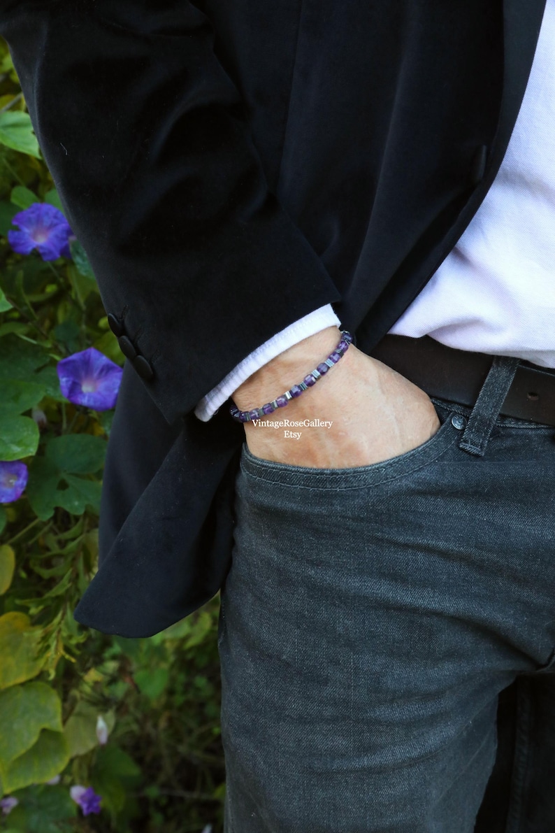 May include: A bracelet with purple and silver beads is worn on a wrist. The person is wearing a white shirt, a black blazer, and blue jeans. The background includes green foliage and purple flowers.