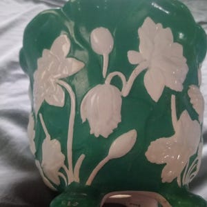 May include: A green ceramic vase with white floral relief detailing. The vase has a glossy finish and a rounded shape, with stylized flowers and leaves on its surface. The design is symmetrical, creating a classic and elegant aesthetic.