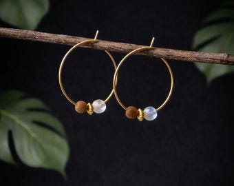 May include: A pair of gold hoop earrings, each adorned with a brown bead and a white, iridescent bead. The earrings are displayed on a thin, brown branch against a dark background, with green leaves in the corners.