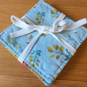 May include: A stack of square fabric coasters tied together with a white cotton ribbon. The top coaster features a light blue background with a repeating pattern of small yellow berries, green leaves, and light green gourds with stitched edges.