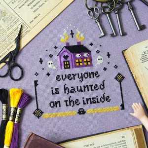 May include: A cross-stitch design on lavender fabric features a purple house with yellow flames and the text "everyone is haunted on the inside." The image includes keys, thread, and vintage ephemera.