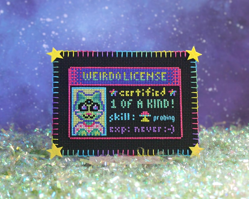 Op de afbeelding: Een kruissteek "WEIRDO LICENSE" met een cartoon kat illustratie. De zwarte achtergrond is omlijst met kleurrijke steken en gele ster accenten. Tekst bevat "certified", "1 of a kind", "skill: probing" en "exp: never :-)".