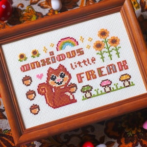 May include: Cross-stitch artwork in a wooden frame. The design features a squirrel, sunflowers, mushrooms, acorns, and a rainbow. Text reads "amawious little FREAK". The color palette includes brown, orange, green, pink, and yellow.