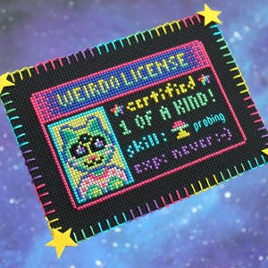 Op de afbeelding: Een kruissteek "WEIRDO LICENSE" met een cartoon kattenportret. De licentie is zwart met kleurrijke steken, met tekst als "certified 1 OF A KIND!" en "skill: probing." Gele steraccenten zijn aan de hoeken bevestigd.