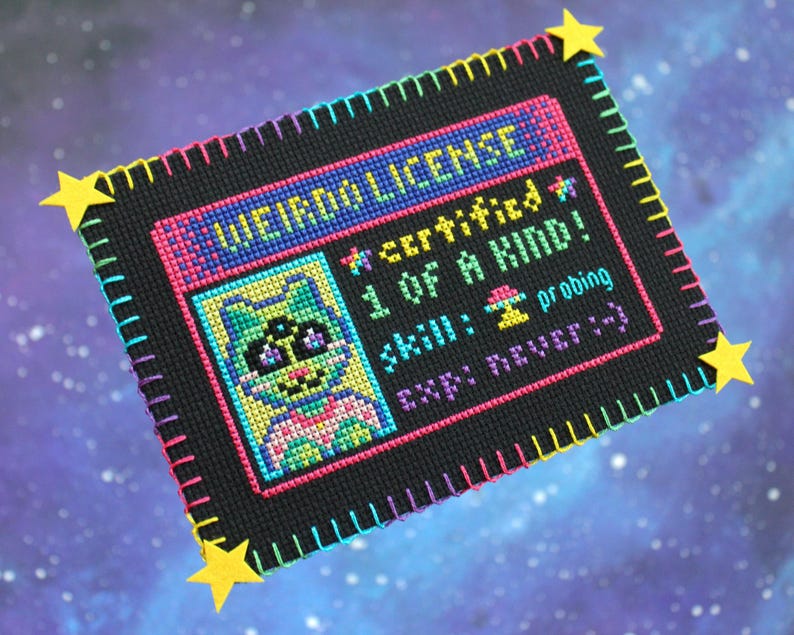 Op de afbeelding: Een kruissteek "WEIRDO LICENSE" met een cartoon kattenportret. De licentie is zwart met kleurrijke steken, met tekst als "certified 1 OF A KIND!" en "skill: probing." Gele steraccenten zijn aan de hoeken bevestigd.