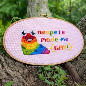 May include: Oval cross-stitch embroidery hoop with a pink background. The design features a colorful cartoon creature with the text "neopets made me GAY!" in rainbow colors. The hoop is made of wood.