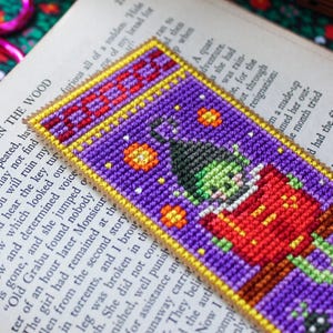 May include: A handmade bookmark with a pixelated witch design. The bookmark is purple with a yellow border and the word "BOO" at the top. The witch wears a red outfit and a green face, ideal for Halloween or fantasy readers.