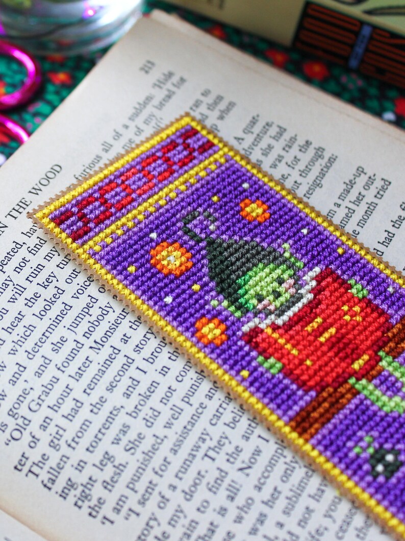 May include: A handmade bookmark with a pixelated witch design. The bookmark is purple with a yellow border and the word "BOO" at the top. The witch wears a red outfit and a green face, ideal for Halloween or fantasy readers.