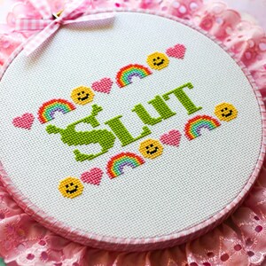 Puede incluir: Un bastidor de bordado con la palabra "SLUT" en verde, rodeada de arco&iacute;ris, corazones y caras sonrientes. El bastidor est&aacute; sobre un fondo de encaje rosa y una cinta de cuadros rosa.