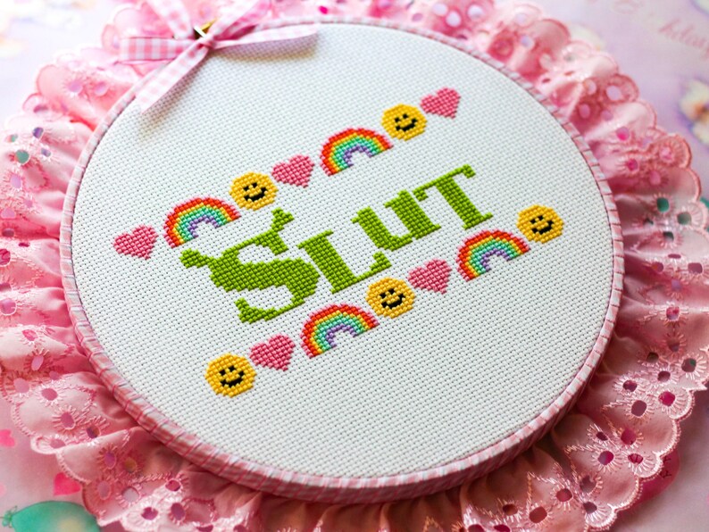 Puede incluir: Un bastidor de bordado con la palabra "SLUT" en verde, rodeada de arco&iacute;ris, corazones y caras sonrientes. El bastidor est&aacute; sobre un fondo de encaje rosa y una cinta de cuadros rosa.