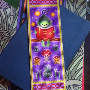 May include: A handmade bookmark with a purple background and a stitched design. The design features a green creature reading a red book, with mushrooms and stars. The bookmark has a yellow border and a black ribbon.